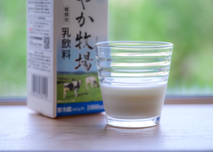 A glass of milk, with a carton of milk and a field in the background.