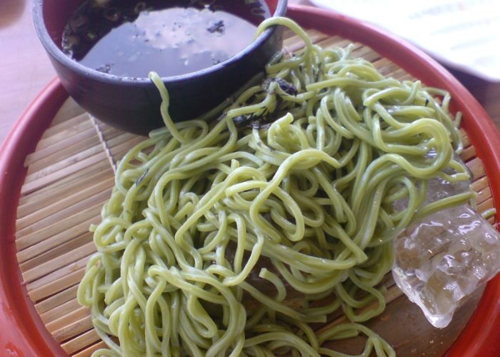 A heap of green soba noodles on a wooden board, accompanied by a dark brown dipping sauce