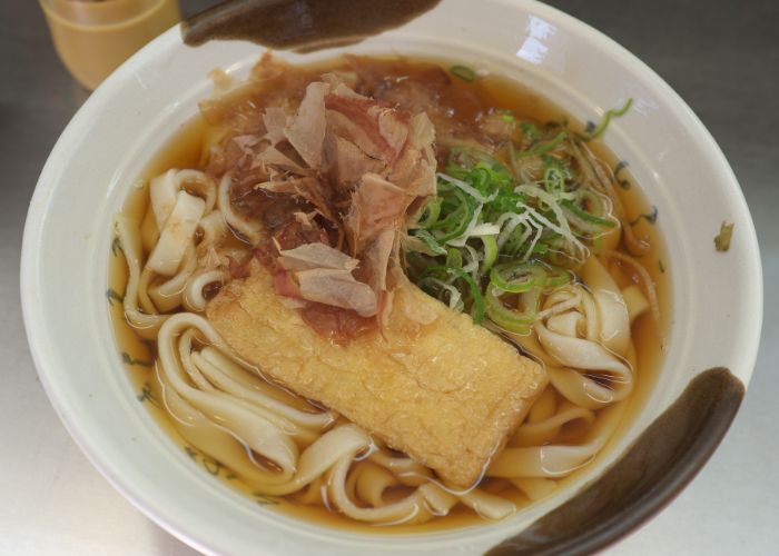 An overhead shot of a bowl of kishimen noodles in a light brown broth, topped with deep-fried tofu, shredded meat and sliced green onions