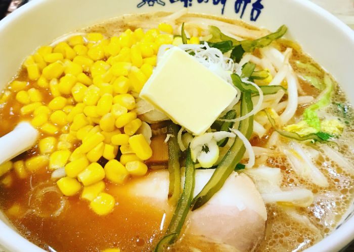 A close-up image of a bowl of Sapporo ramen, with bright yellow sweetcorn, a pat of butter, and noodles in a light brown broth