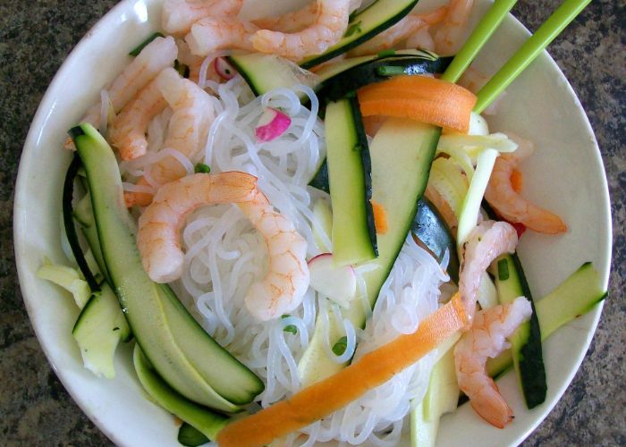 An overhead image of a bowl of white shirataki noodles, topped with sliced carrot, sliced courgette and prawns