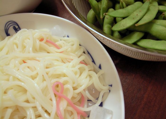 A bowl of white and pink hiyamugi noodles in the foreground, with a plate of edamame behind 