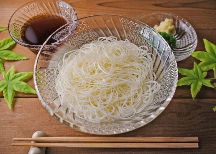 A glass bowl of thin white somen noodles, with glass bowls of dipping sauce and garnishes behind it and wooden chopsticks in front