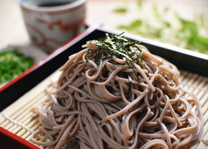 A pile of brown soba noodles on a bamboo tray, topped with thin slices of dry seaweed