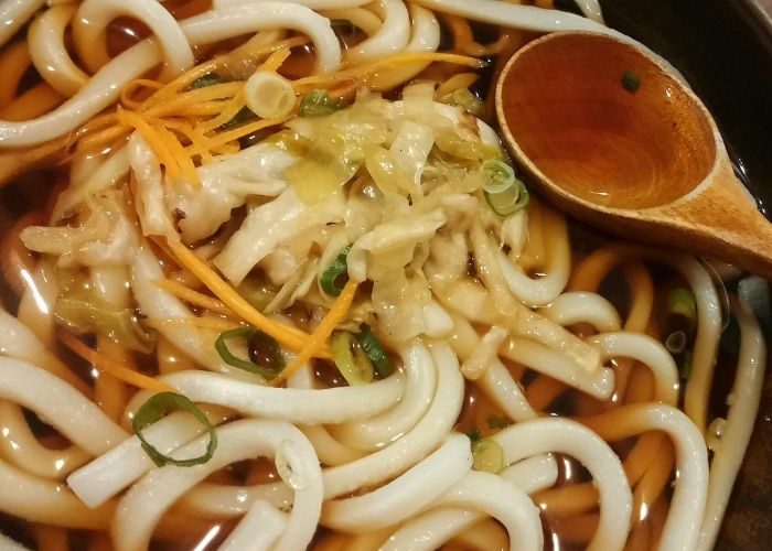 A close up image of a bowl of udon noodles in a light brown broth and topped with vegetables, with a wooden spoon resting on top