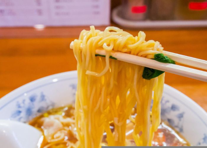 A pair of wooden chopsticks lifting thin yellow ramen noodles out of a bowl