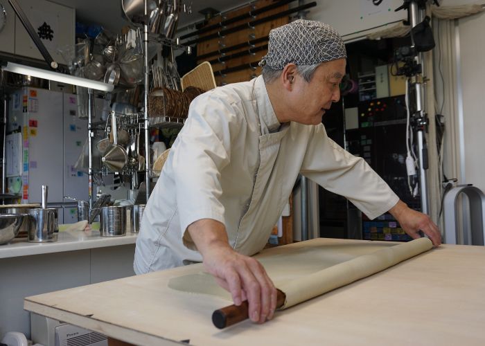 A photo of a male chef in a kitchen, rolling out dough ready to be cut into soba noodles