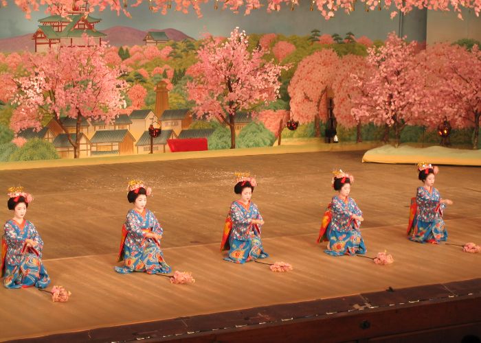 Five geisha kneeling on a stage as part of a performance, with sakura trees in the background