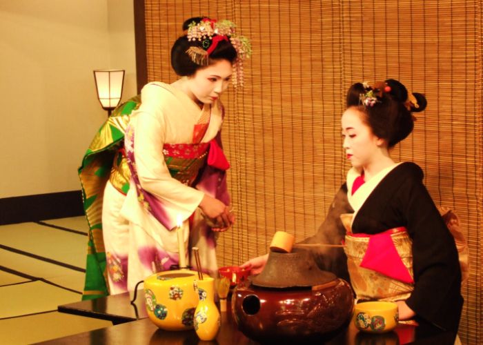 Two geisha in kimono making preparations for a tea ceremony in a tatami mat room