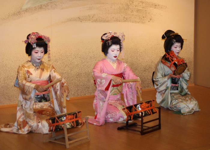 Three geisha kneeling in a row playing traditional Japanese drums