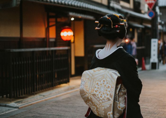 A photo of a geisha walking away from the camera, with a traditional Japanese building in the background 