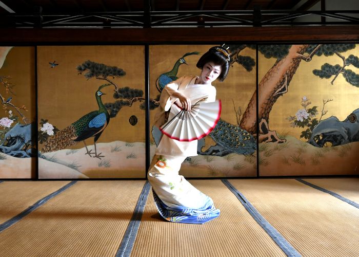 A solo geisha in a white kimono dancing with a red-edged fan in a traditional tatami mat room