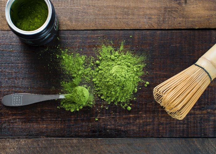 An overhead image of bright green matcha powder on a wooden surface, next to a bamboo whisk, little metal spoon and pot of matcha powder