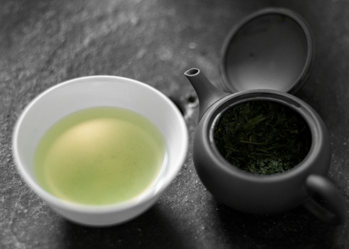 A close up image of a small black teapot with its lid off and dark green tea leaves inside, next to a small white teacup with light green tea inside