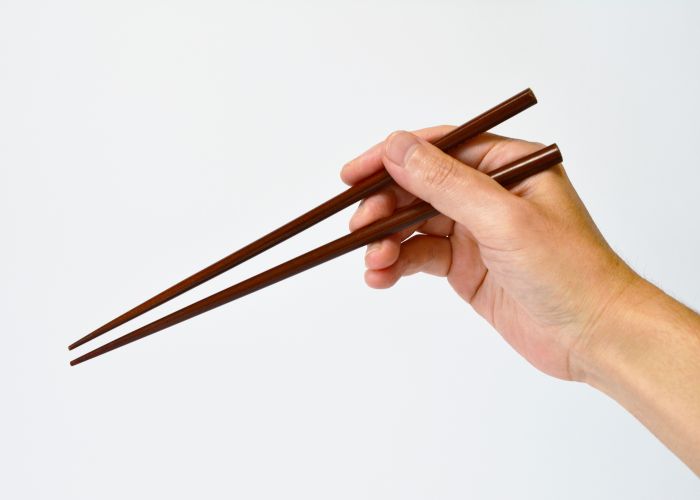 An image of a hand holding a pair of dark brown wooden chopsticks against a white background
