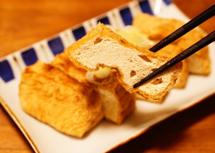 An image of a pair of black chopsticks holding a piece of sliced deep-fried tofu up to the camera, with the remaining slices on a plate in the background