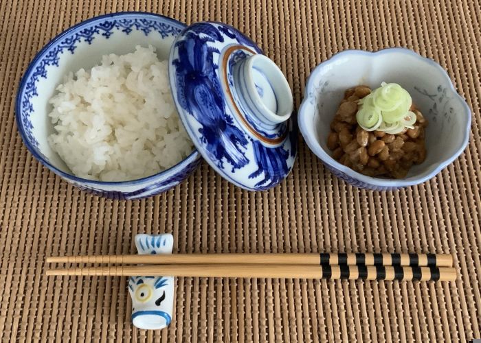A pair of chopsticks laid horizontally in the foreground on a fish-shaped chopstick holder, with a bowl of rice and bowl of natto behind