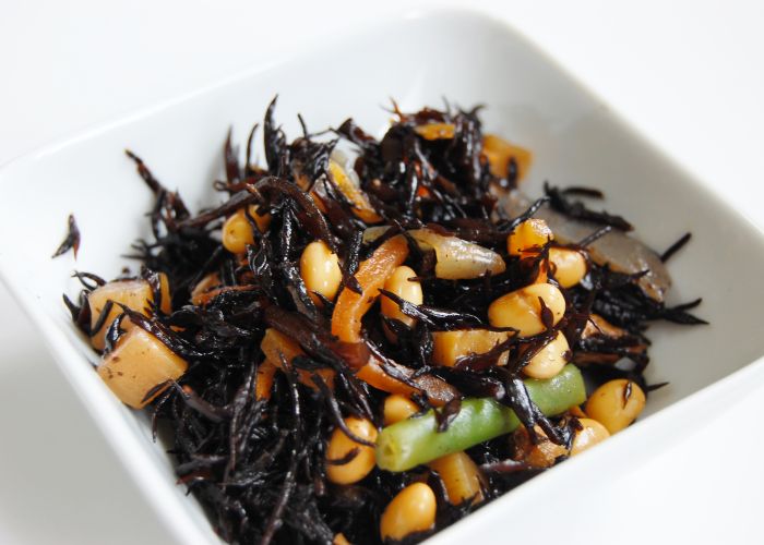 A close up image of a salad in a white bowl, with thin strands of dark brown hijiki seaweed and other vegetables