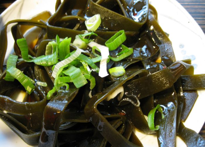 A close up of long, thick strands of dark green seaweed topped with chopped spring onion