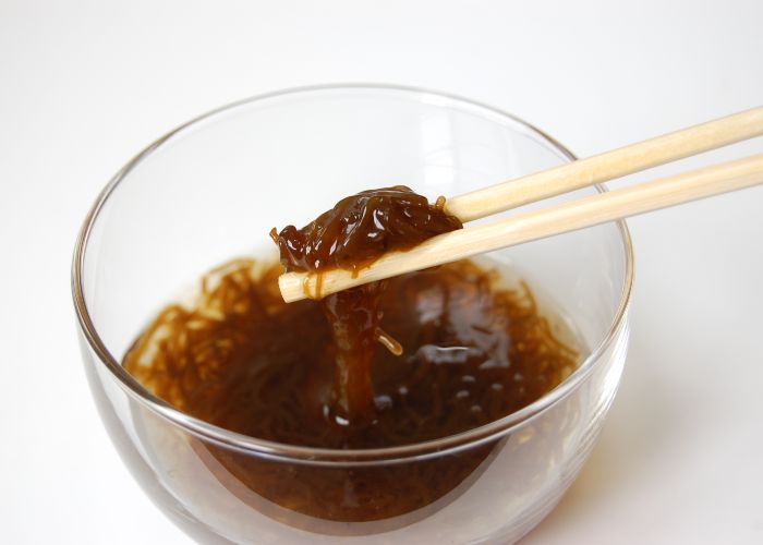 A close up image of a glass bowl full of light brown mozuku seaweed, with a pair of wooden chopsticks lifting some up