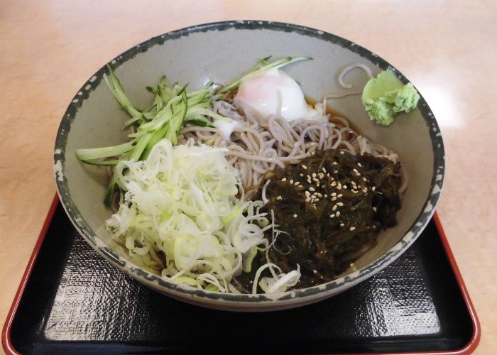 An overhead image of a bowl of soba noodles, topped with mekabu seaweed with sesame seeds, cucumber and spring onions