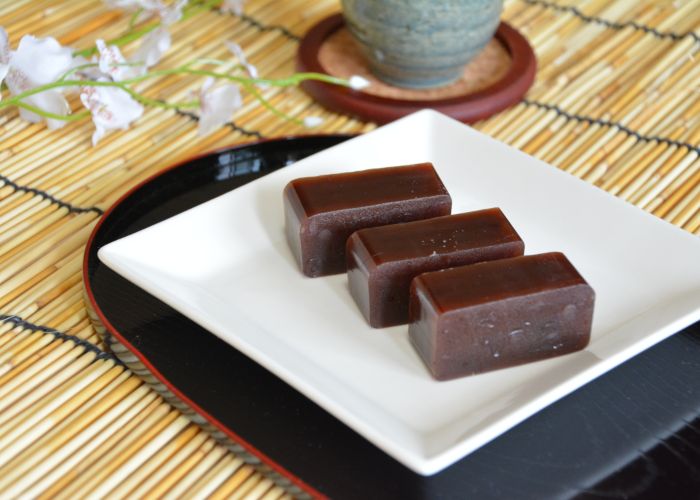 Three rectangles of red yokan sweets on a square white dish, with flowers in the background