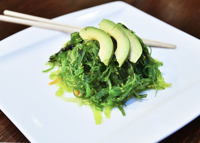 Bright green seaweed salad topped with three slices of avocado, on a white plate with a pair of chopsticks