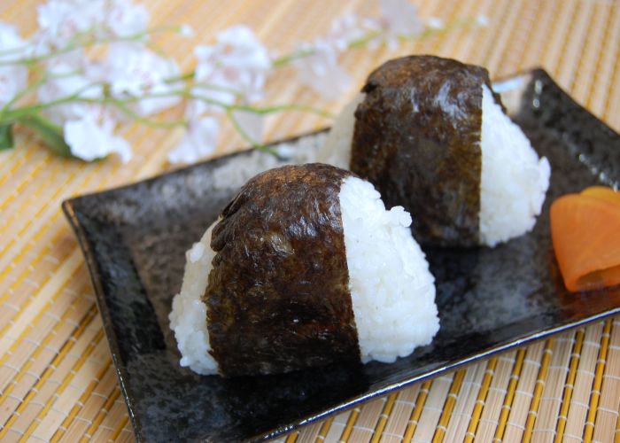 Two onigiri rice balls wrapped in a strip of nori seaweed, on a black plate with flowers in the background