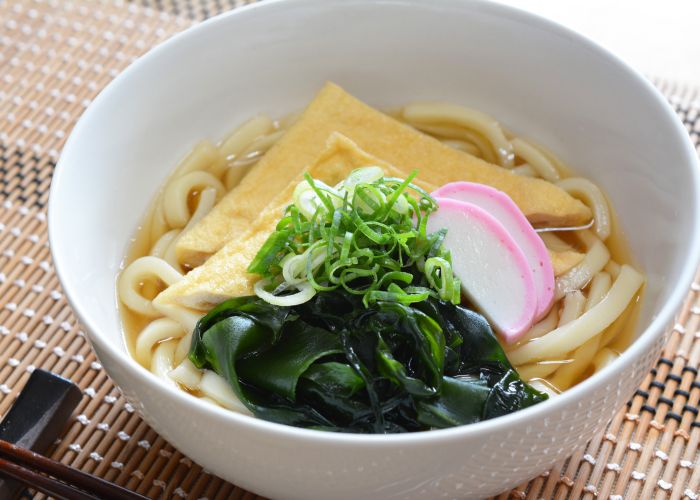 A bowl of udon noodles, topped with a pile of seaweed, tofu and spring onions