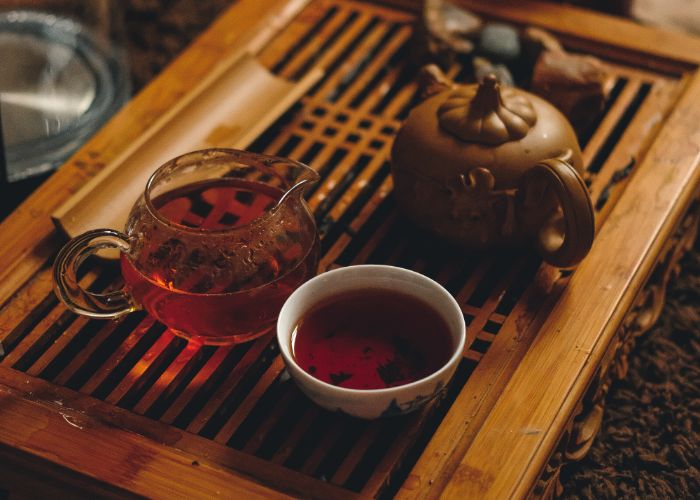 A clay teapot, glass tea jug and ceramic teacup filled with brown tea on a wooden tray