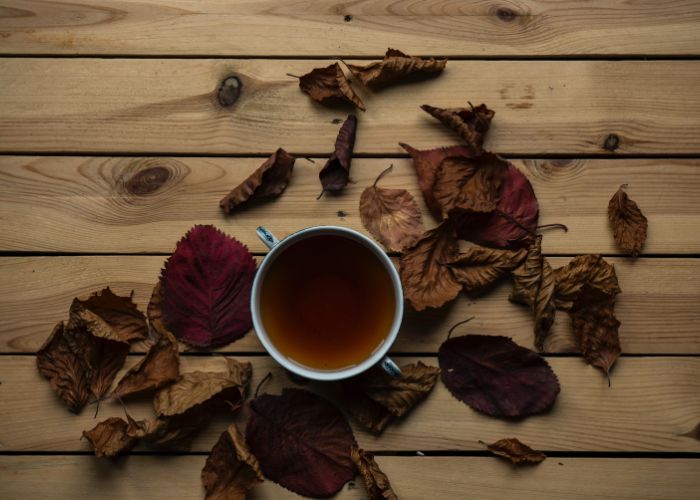 An overhead image of a cup of brown tea, surrounded by autumn leaves on a wooden surface