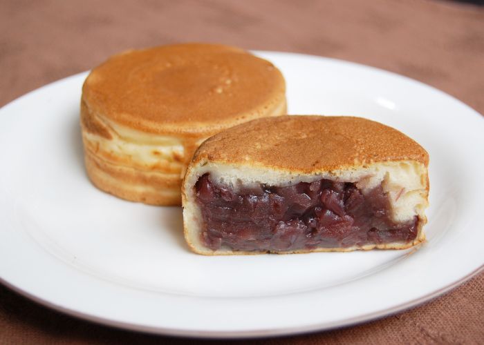 Two round golden imagawayaki on a white plate, one cut in half to reveal a thick filling of red bean paste