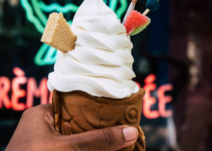 A close up of a hand holding a taiyaki cone filled with white soft serve ice cream and topped with a wafer