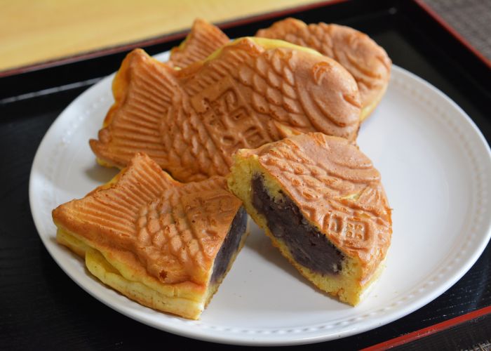 Three fish-shaped taiyaki on a white plate, with the front one cut in half to reveal a thick filling of anko