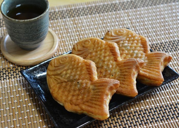 Three golden fish-shaped taiyaki lined up on a black rectangular plate on top of a mat, with a tea cup in the background