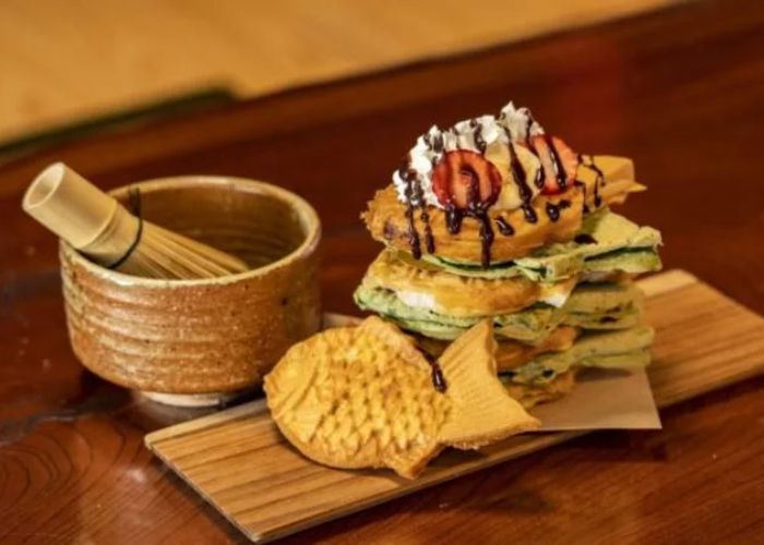 A stack of taiyaki, topped with fruit, whipped cream and chocolate sauce, with a matcha whisk and bowl in the background