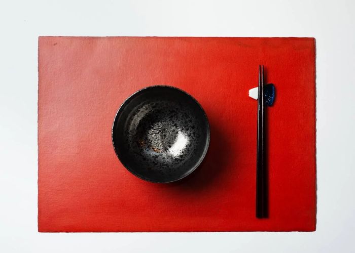 An overhead image of a black bowl beside a pair of black chopsticks, all on a red tablemat