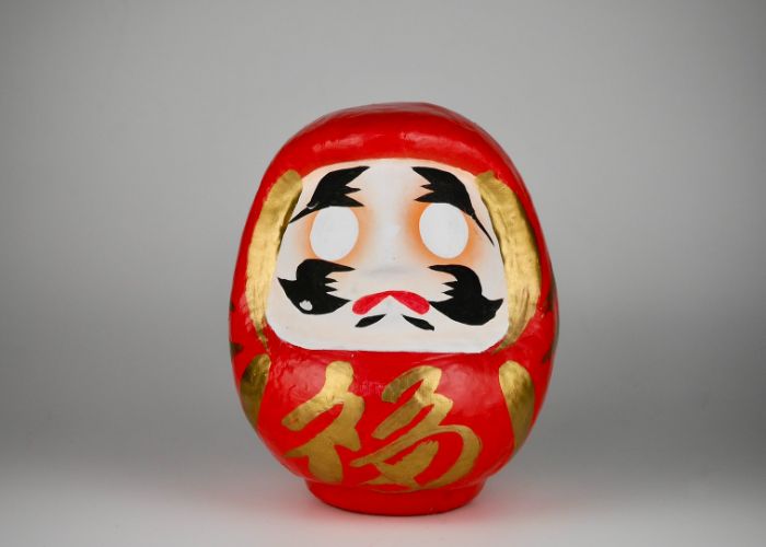 A round red, black and gold daruma doll with two blank white eyes