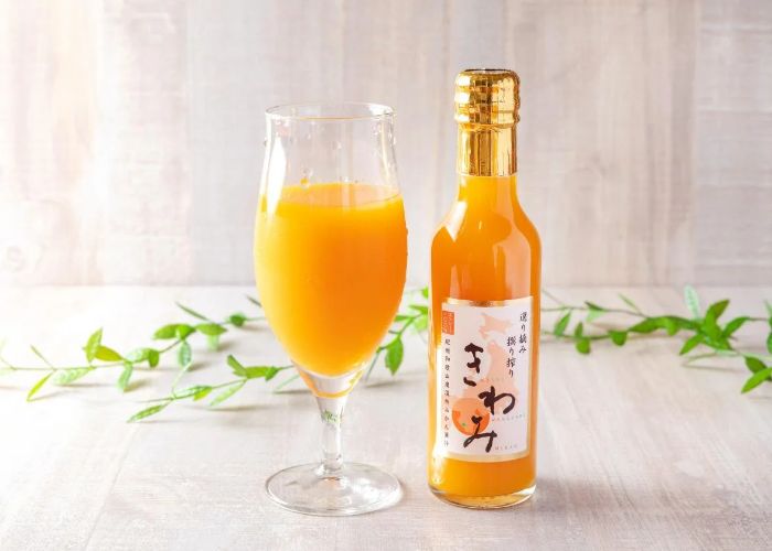 A full bottle of mikan fruit juice next to a glass full of orange mikan juice, with a green leafy branch in the background