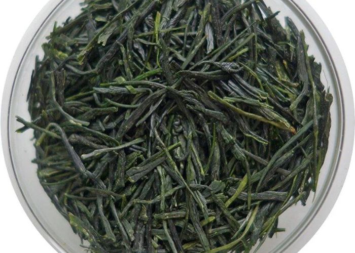 A close up overhead shot of a glass bowl full of dried, long, thin green tea leaves
