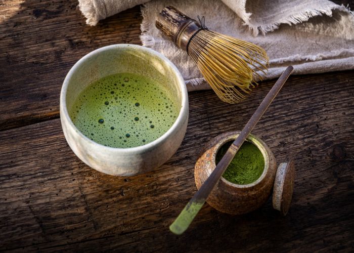 A beige bowl of bright green frothy matcha tea, next to a whisk and a pot of green matcha powder