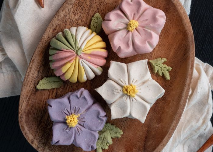 An overhead close-up image of four wagashi sweets on a wooden plate: a pink flower, a white flower, a purple flower and a round multicolored one