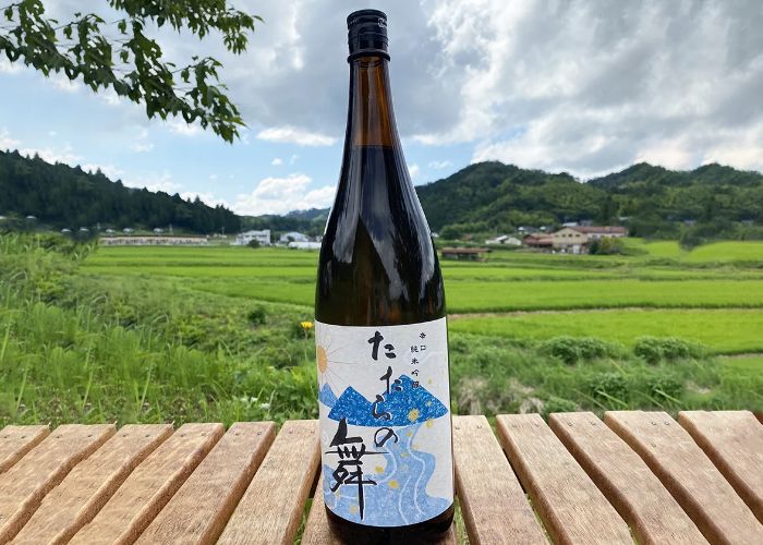 A brown bottle of sake with a blue and white label on wooden decking with green fields in the background