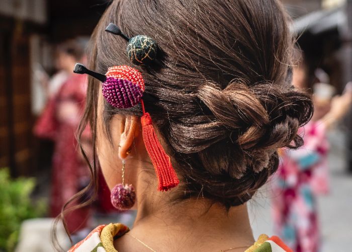 A close up photo of the back of a woman's head, with two round Japanese hair accessories in her hair, one purple and red with a red tassel, and the other green and gold