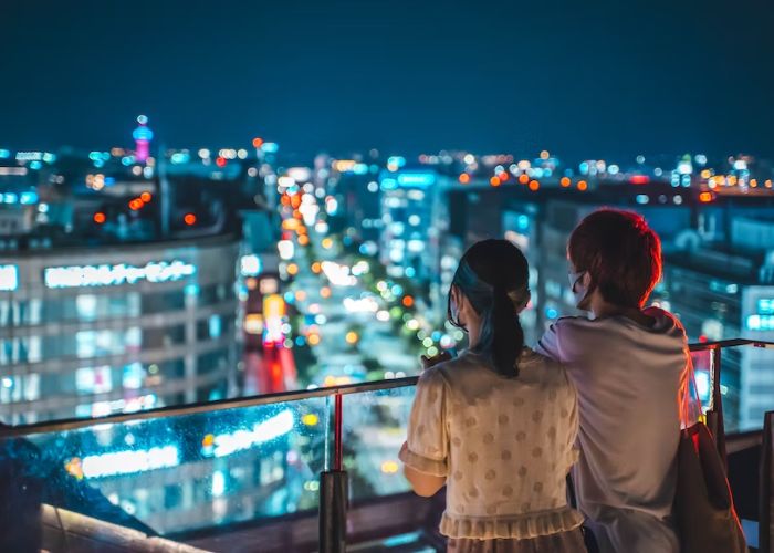 A Japanese couple with their backs to the camera overlook a night-time cityscape from a highrise balcony in Japan.