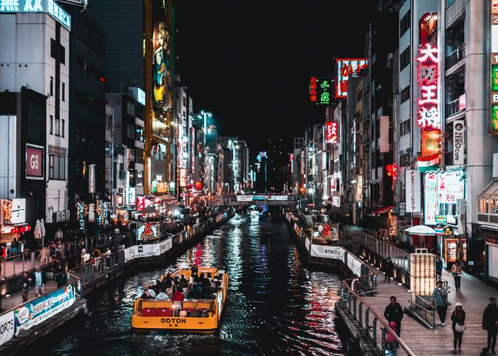 A photo of Dotonburi at night, with a boat traveling down the river in the center and buildings with neon lights on either side