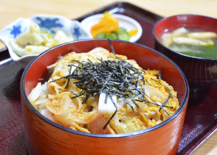 A circular wooden box filled with oyakodon donburi, topped with shredded seaweed. In the background there is also a soup and side dishes on the tray.