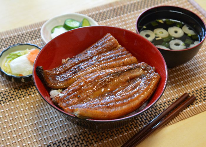 A wooden bowl of white rice topped with slices of grilled eel, with miso soup and pickles in the background