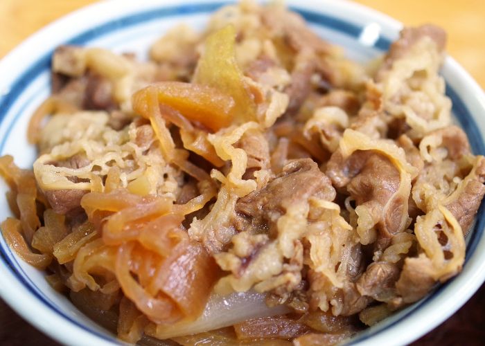 A close up image of a white bowl of gyudon, topped with thin strips of cooked onion and beef