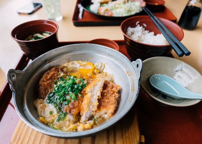 A tray of food with a bowl of katsudon in the front, topped with a sliced breaded cutlet and green onions. In the background is a bowl of soup and separate dish of rice